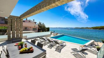 Piscina en la azotea con sillones de descanso y una mesa con fruta, con vistas a un mar azul y una costa lejana.