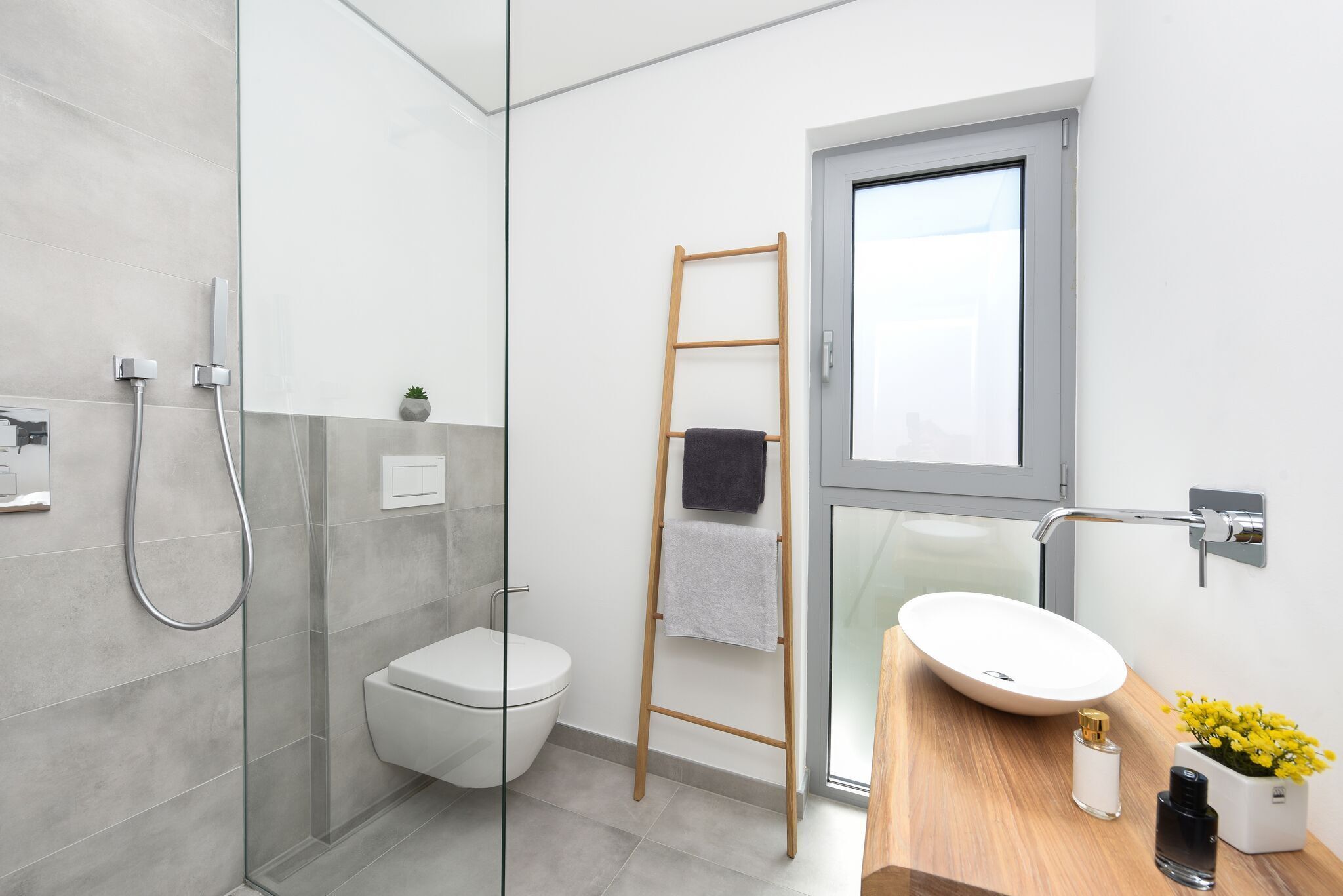 Un baño moderno con cabina de ducha de vidrio, inodoro blanco, escalera de madera con una toalla, lavamanos blanco sobre encimera de madera y una pequeña planta en maceta.