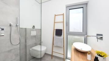 Un baño moderno con cabina de ducha de vidrio, inodoro blanco, escalera de madera con una toalla, lavamanos blanco sobre encimera de madera y una pequeña planta en maceta.