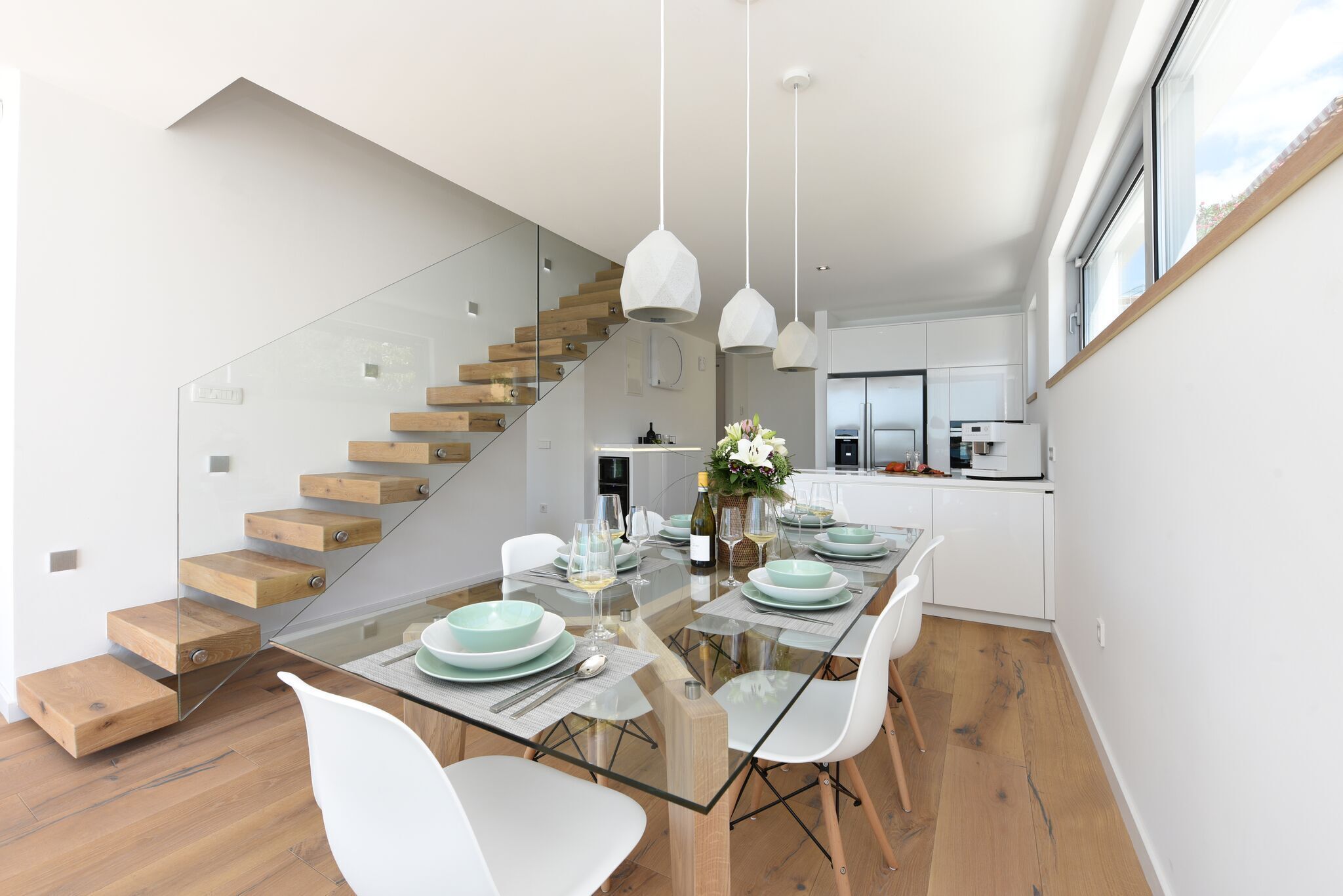 Una cocina moderna con armarios blancos, una escalera de cristal y un comedor con una mesa de cristal y sillas blancas.