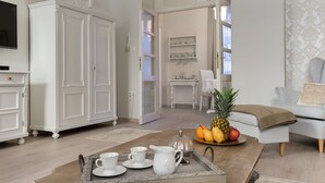 A spacious living room with a white cabinet, a coffee table with a tray of white cups and a pitcher, a fruit bowl with pineapple and oranges, and a grey sofa with a beige throw blanket. - Chestnut & Eliza Suites - Superior Homes (Budapest)