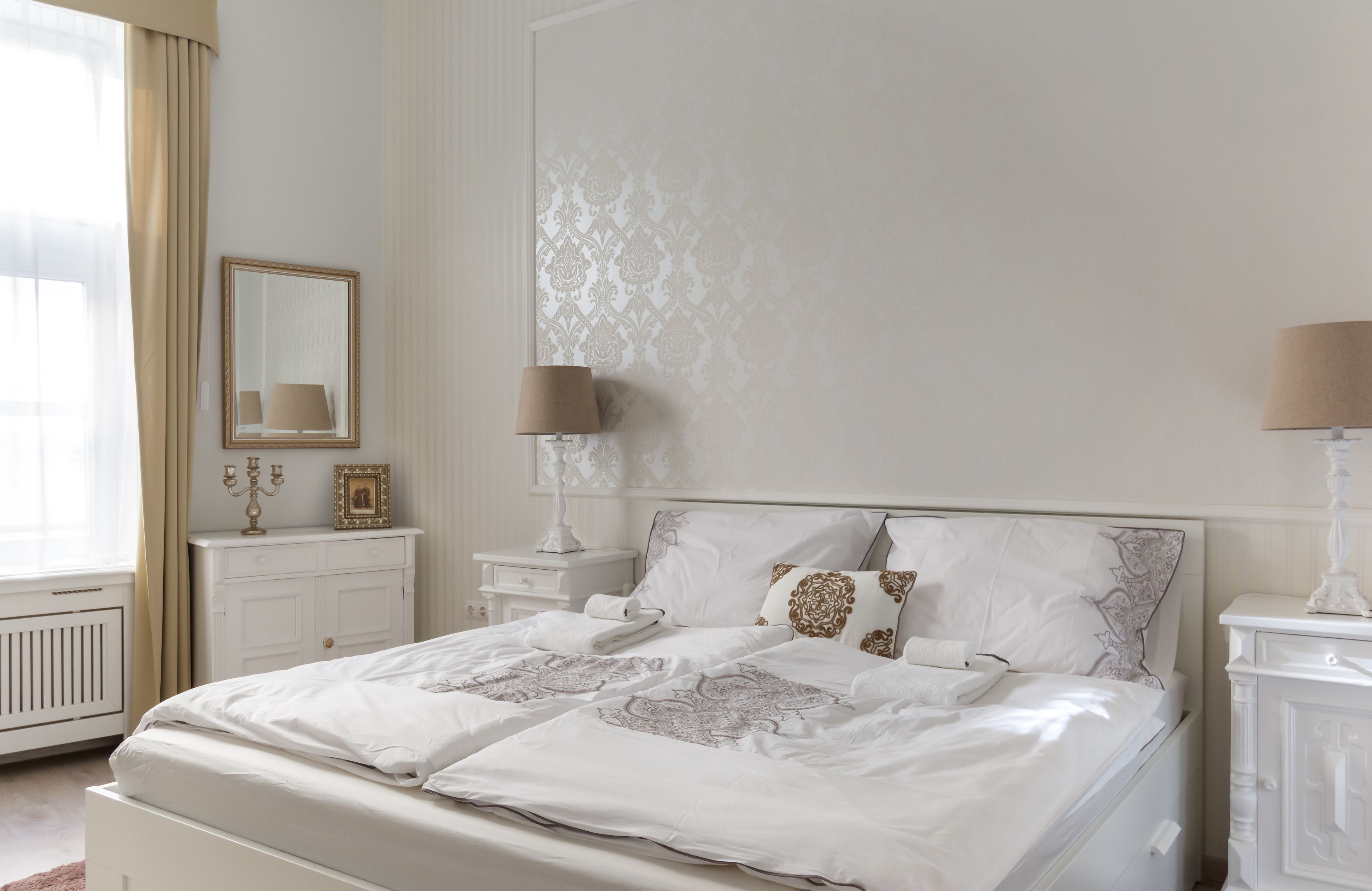 Un dormitorio con una cama grande, dos mesitas de noche, un tocador, un espejo y una ventana con cortinas.