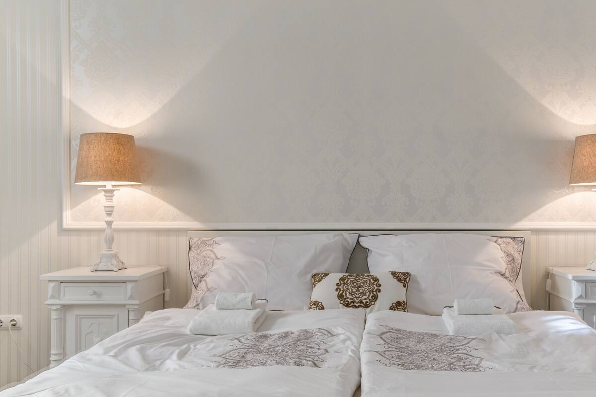 Een keurig opgemaakt bed met witte lakens en gestreepte kussens, met aan beide kanten nachtkastjes met lampen.
