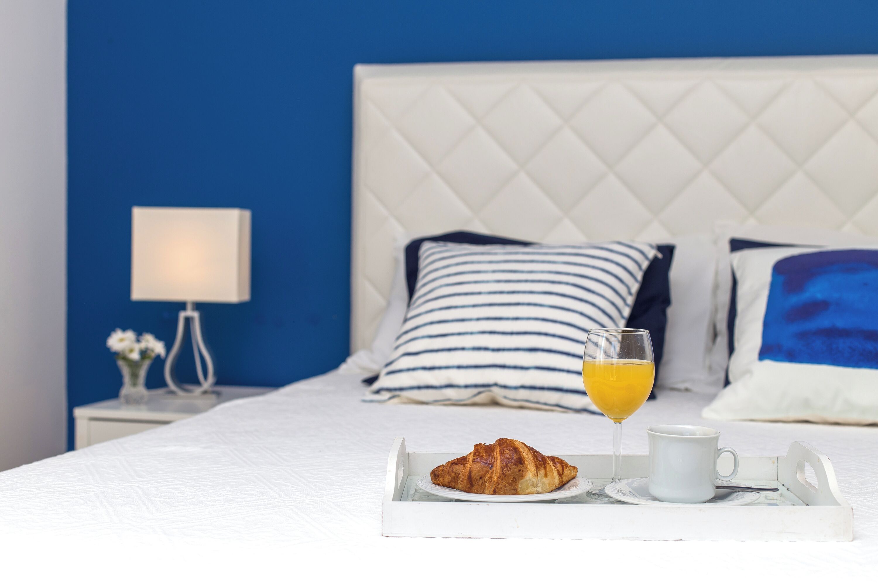 A neatly made bed with a white quilt, a bedside lamp, and striped pillows. A tray with a croissant, a glass of orange juice, and a white mug is placed on the bed.