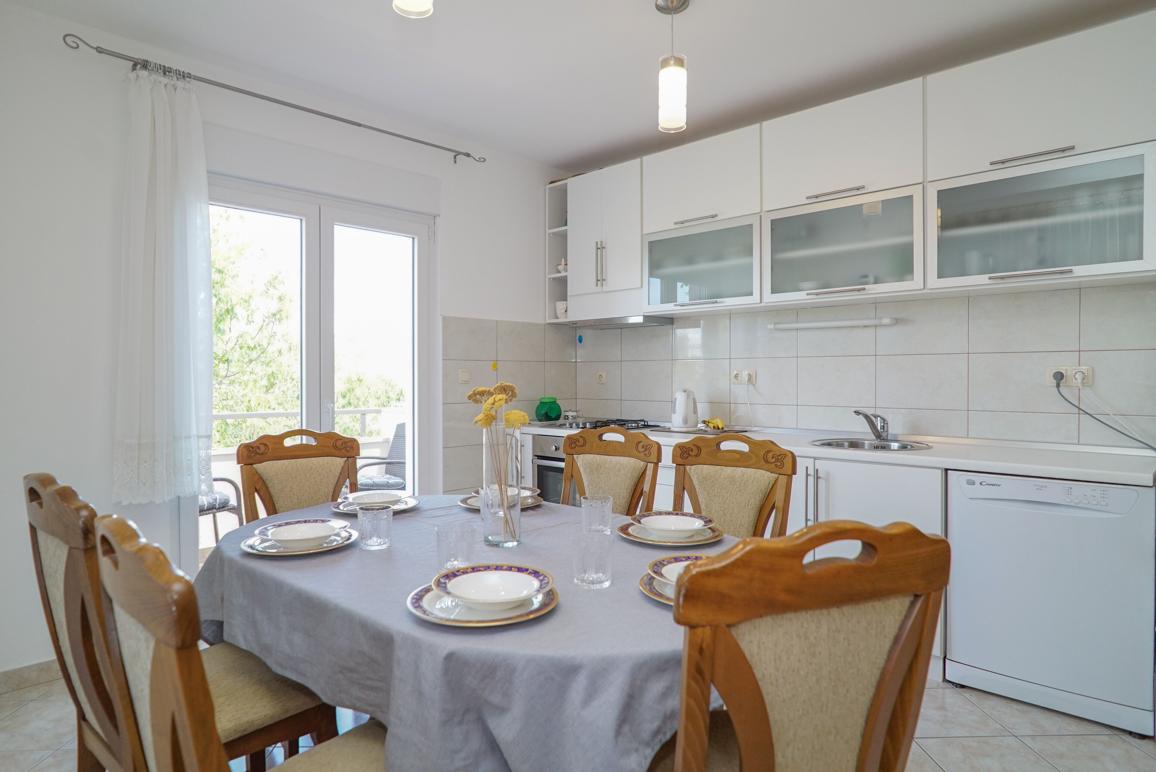 A dining area with a round table set for six, surrounded by wooden chairs. A kitchen with white cabinets and a dishwasher is visible in the background.
