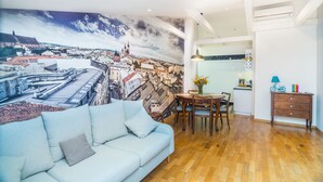 Uma sala de estar moderna com um grande mural de paisagem urbana, um sofá azul claro, piso de madeira, uma área de jantar com mesa e cadeiras, e uma cozinha compacta ao fundo.
