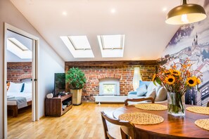 A cozy living room with a brick fireplace, a dining table with placemats, and a large window with skylights.
