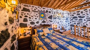A bedroom with a stone wall, wooden ceiling beams, a bed with floral bedding, and wall-mounted lights. - Adega Velha holiday home, next to the sea and mountains, ideal for couples (Calheta de Nesquim)