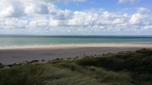 A beach with a wide sandy shore, grassy dunes in the foreground, and a calm sea under a partly cloudy sky.