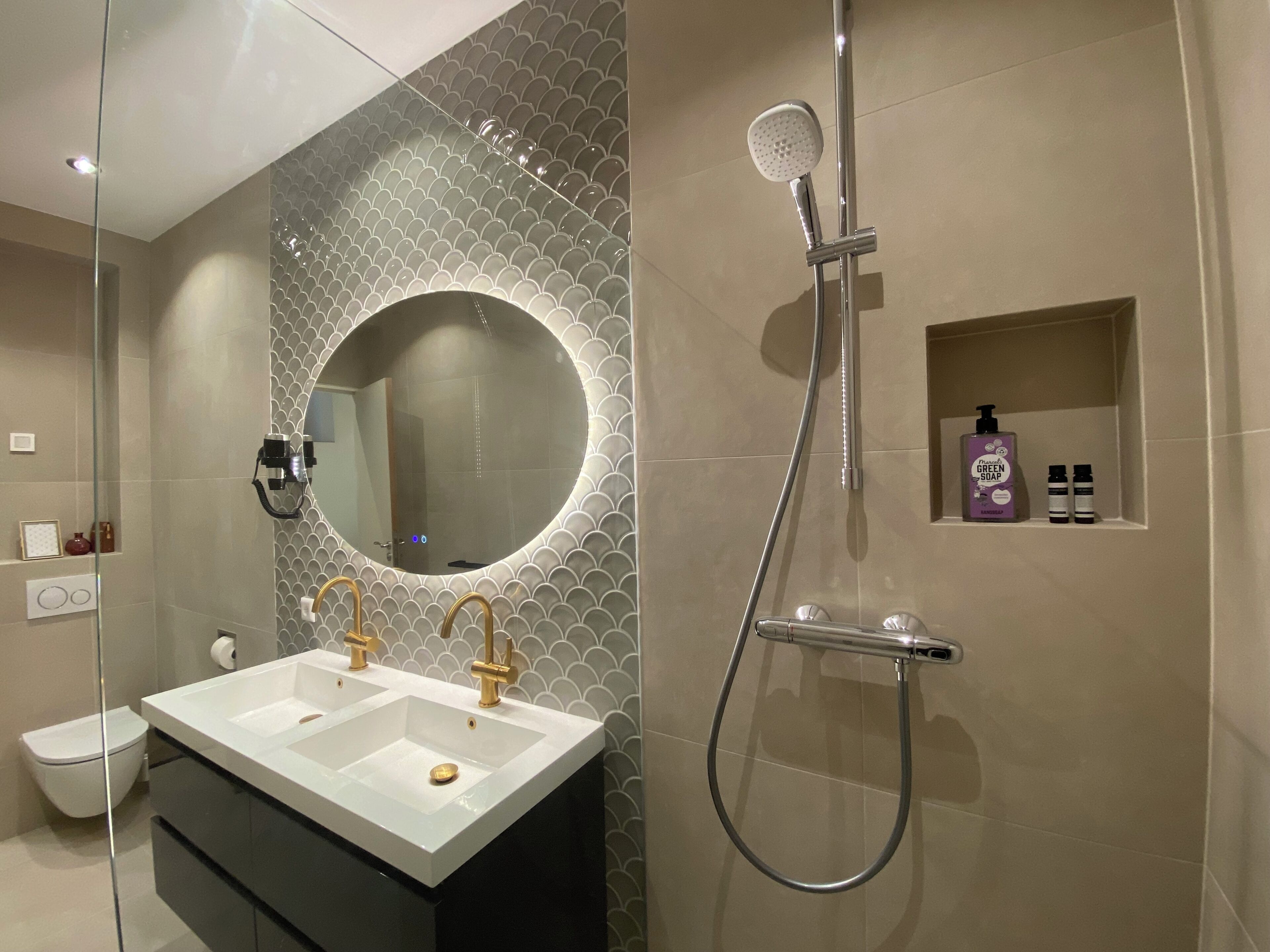 A bathroom with a double sink, a shower, a mirror, and a shelf with toiletries.