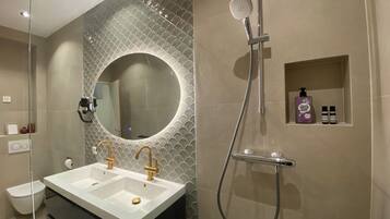 A bathroom with a double sink, a shower, a mirror, and a shelf with toiletries.