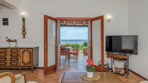 Een woonkamer met een televisie, houten meubels, een eettafel en uitzicht op zee door een open deur.