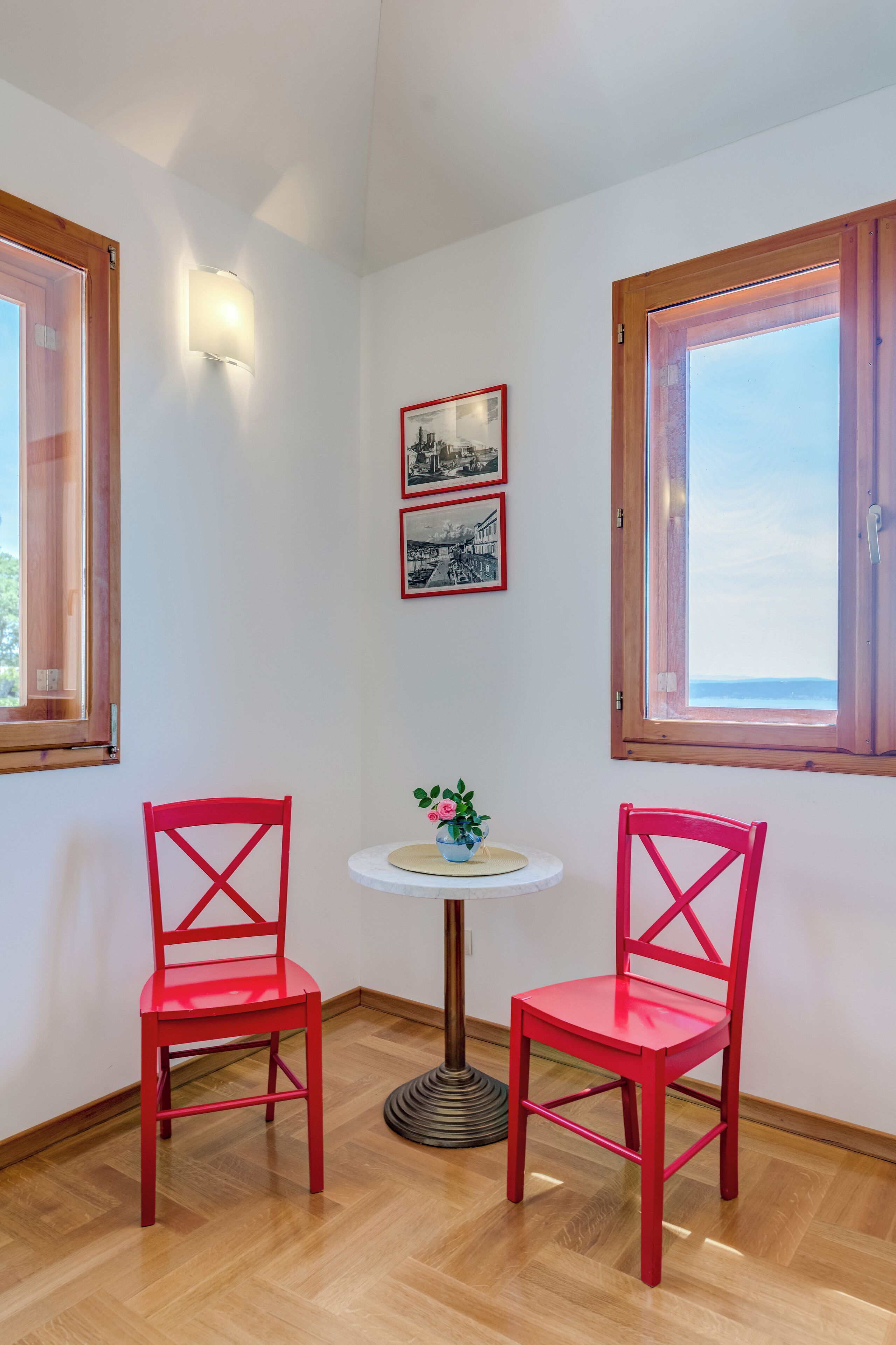 Une pièce d’angle avec deux fauteuils rouges, une petite table ronde ornée d’un pot de fleurs et des tableaux encadrés accrochés au mur.