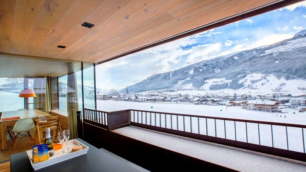 Une pièce avec un plafond en bois, une table à manger avec des chaises, un mur en verre et une vue sur un village enneigé entouré de montagnes.