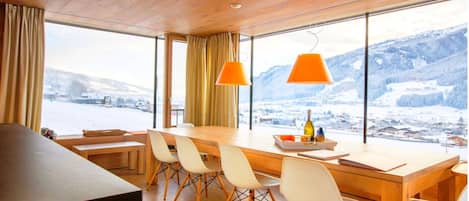A modern dining area with a wooden table, white chairs, and large windows offering a view of a snowy landscape.