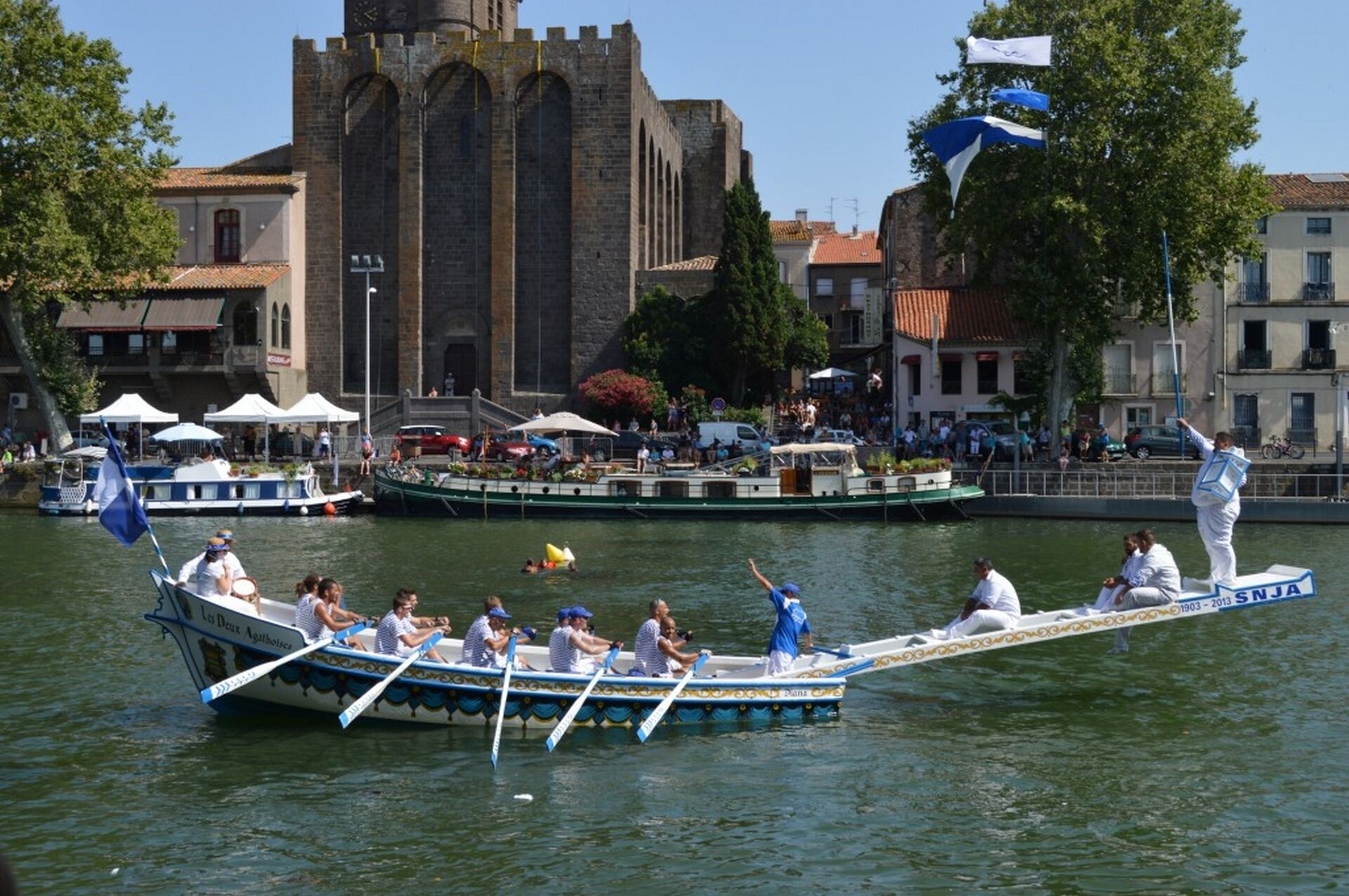 Ein traditionelles Bootsrennen mit Teilnehmern in einem schmuckvollen, schmalen Boot auf einem Fluss, Zuschauern an den Ufern und einem historischen Gebäude im Hintergrund.