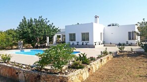 A modern white building with a swimming pool, surrounded by greenery and stone walls.