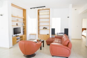 A modern living room with a sectional sofa, a flat-screen TV, wooden shelving, and a dining area.