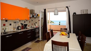 Uma cozinha moderna com armários laranja, uma área de jantar com mesa posta para dois e vista para o mar pela janela.