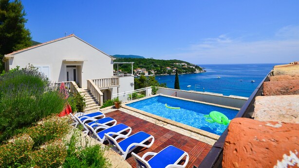 Uma área de piscina com cadeiras de lounge azuis, um pequeno edifício e vista para o mar.