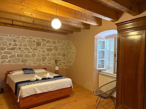 A bedroom with a stone wall, wooden ceiling beams, a bed with a white and blue bedspread, a chair, and a wooden wardrobe.