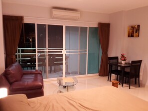 Una habitación de hotel moderna con un gran ventanal corredero que da a un balcón, un sofá de cuero, una pequeña mesa redonda y un comedor con sillas y un jarrón de flores.