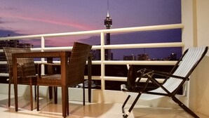 Ein Balkon mit Korbstühlen und einem schwarzen Klappstuhl mit Blick auf eine Stadtlandschaft, dominiert von einem markanten Turm.