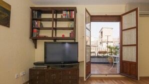 Una sala de estar con televisor, mueble de madera y puerta abierta que da a un balcón con mobiliario exterior.