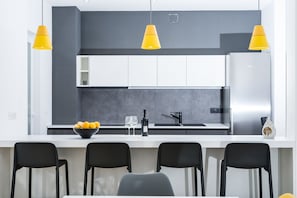 A modern kitchen with white cabinets, a black countertop, and a stainless steel refrigerator. There are three yellow pendant lights, a bowl of fruit, and two wine glasses on the counter. - Comfortable Villa with private pool and sauna near Zadar, Croatia (Privlaka)