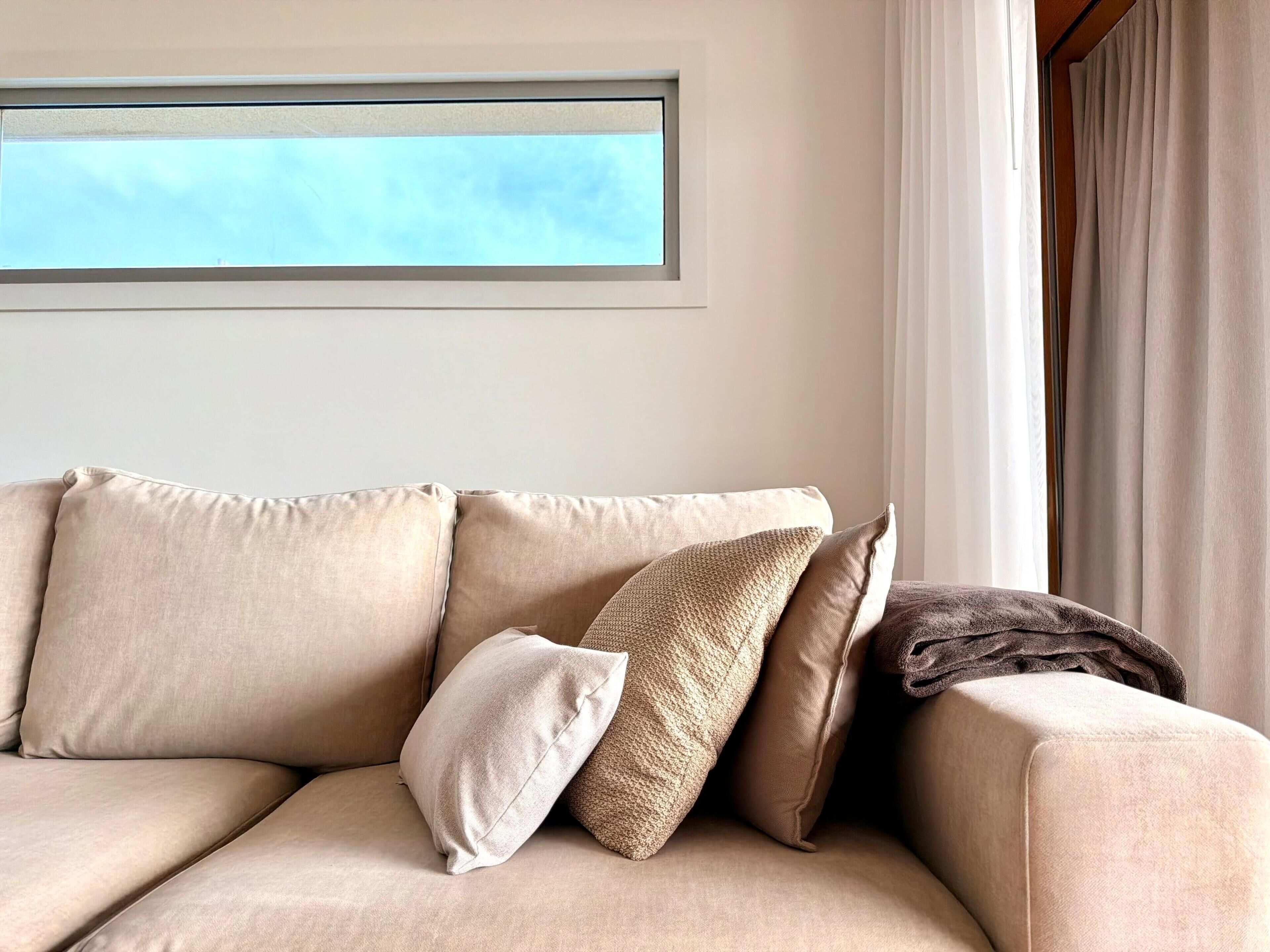 A beige sofa with multiple cushions in a room with a window and curtains.