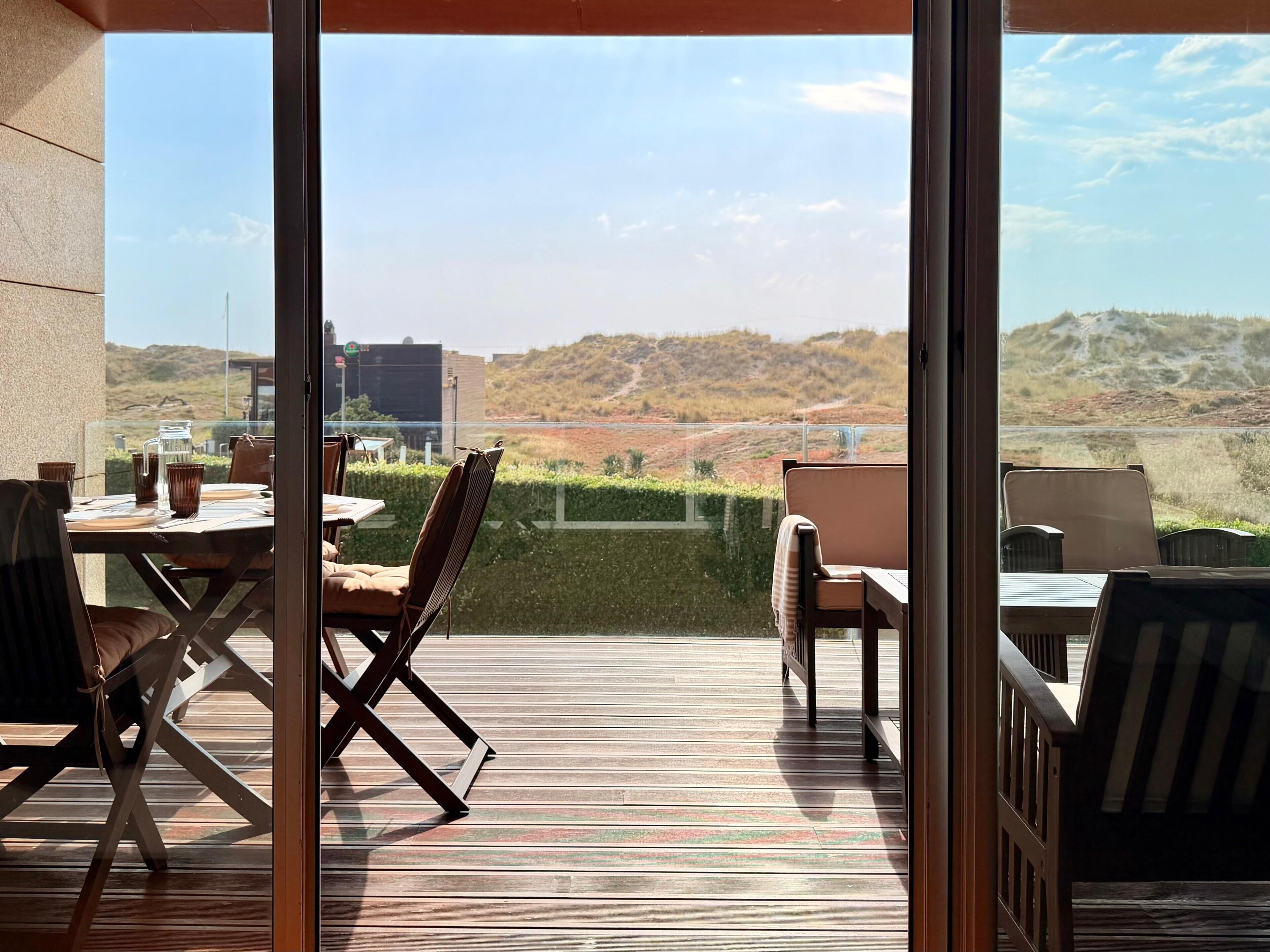 En udendørs terrasse med et bord og stole, samt en glasdør der leder til udsigt over et kuperet landskab.
