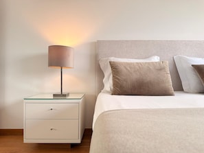 Un letto rifatto con biancheria bianca e beige, una lampada da comodino e un piccolo comodino bianco con cassetti.