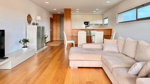 Een moderne woonkamer met een hoekbank, een flatscreen televisie, een houten vloer en een keuken met een eettafel en stoelen.