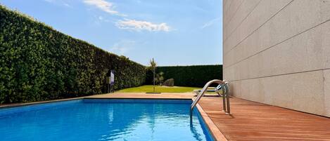A swimming pool with a wooden deck, a hedge, and a modern building in the background.