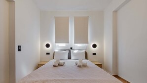 Una cama bien hecha con dos almohadas y dos toallas enrolladas, una mesita de noche y dos luces fijadas a la pared.