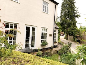 Une maison à deux étages avec un jardin à l’avant, un petit patio avec une table et des chaises, et un grand arbre sur la droite.
