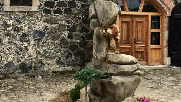 Eine Steinstatue eines Bären, der auf Steinen balanciert, vor einem Gebäude mit einer Steinmauer und einer Holztür.