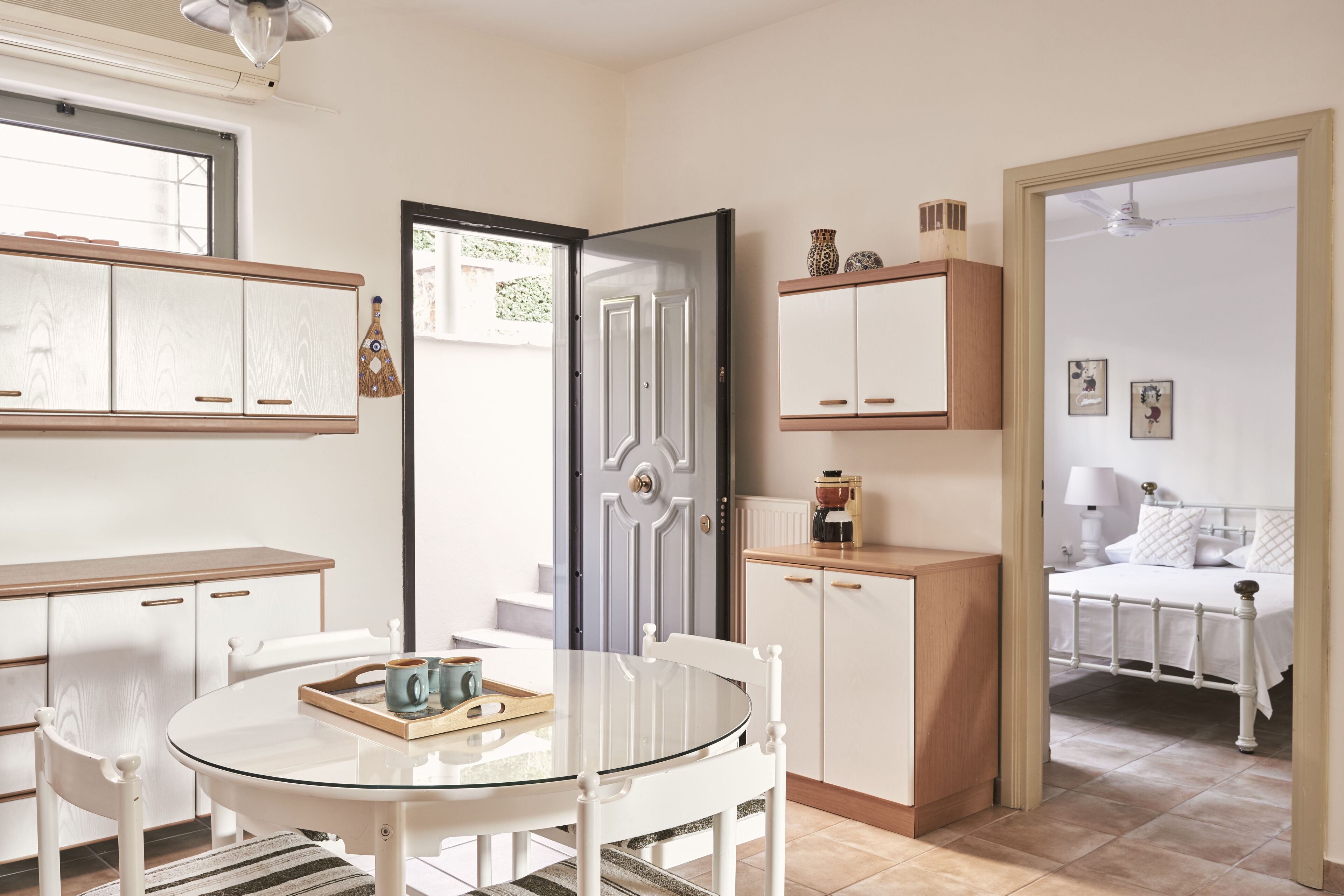 Quarto compacto e bem iluminado com uma mesa de jantar, cadeiras, cozinha com armários e uma cama no quarto adjacente.