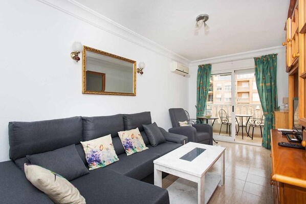 Una sala moderna con un sofá gris, una mesita de centro blanca y un espejo grande. Cuenta con un balcón con mesa y sillas, y un mueble de madera con un televisor.