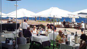 Un espacio de comedor al aire libre con mesas y sillas, sombrillas blancas y vistas al mar.