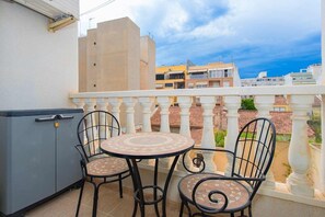 Un balcón con una mesa redonda y dos sillas, un armario gris, y vistas a edificios en el fondo.