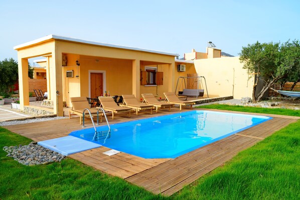 A pool area with a wooden deck, lounge chairs, and a small building with a door and windows.