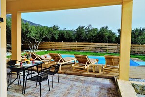 An outdoor patio with a pool, wooden lounge chairs, a dining table with chairs, and a view of a mountain and greenery.