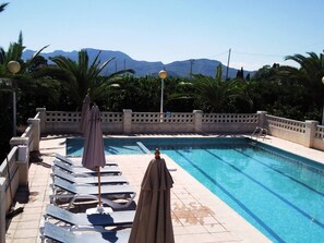 A swimming pool with lounge chairs and umbrellas, surrounded by a tiled deck and palm trees.