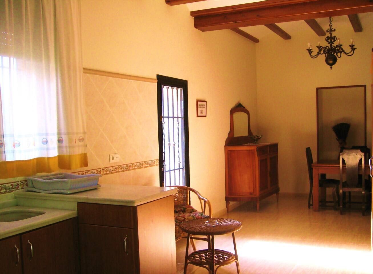 Een kamer met een keukenhoek, een eettafel met stoelen, een kroonluchter en een raam met gordijnen.