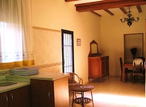 A room with a kitchen area, a dining table with chairs, a chandelier, and a window with curtains. - Valencia Country Hotel - Villamilagro (Polinyà de Xúquer)