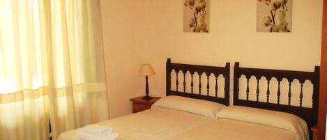Una cama doble con cabecera de madera, una mesita de noche con lámpara, y dos cuadros de algodón colgados en la pared.