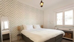 Un dormitorio con cama, espejo, lámpara y ventana.