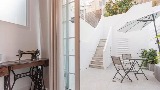Una habitación con una máquina de coser, una mesa con sillas, una escalera y vistas al exterior.