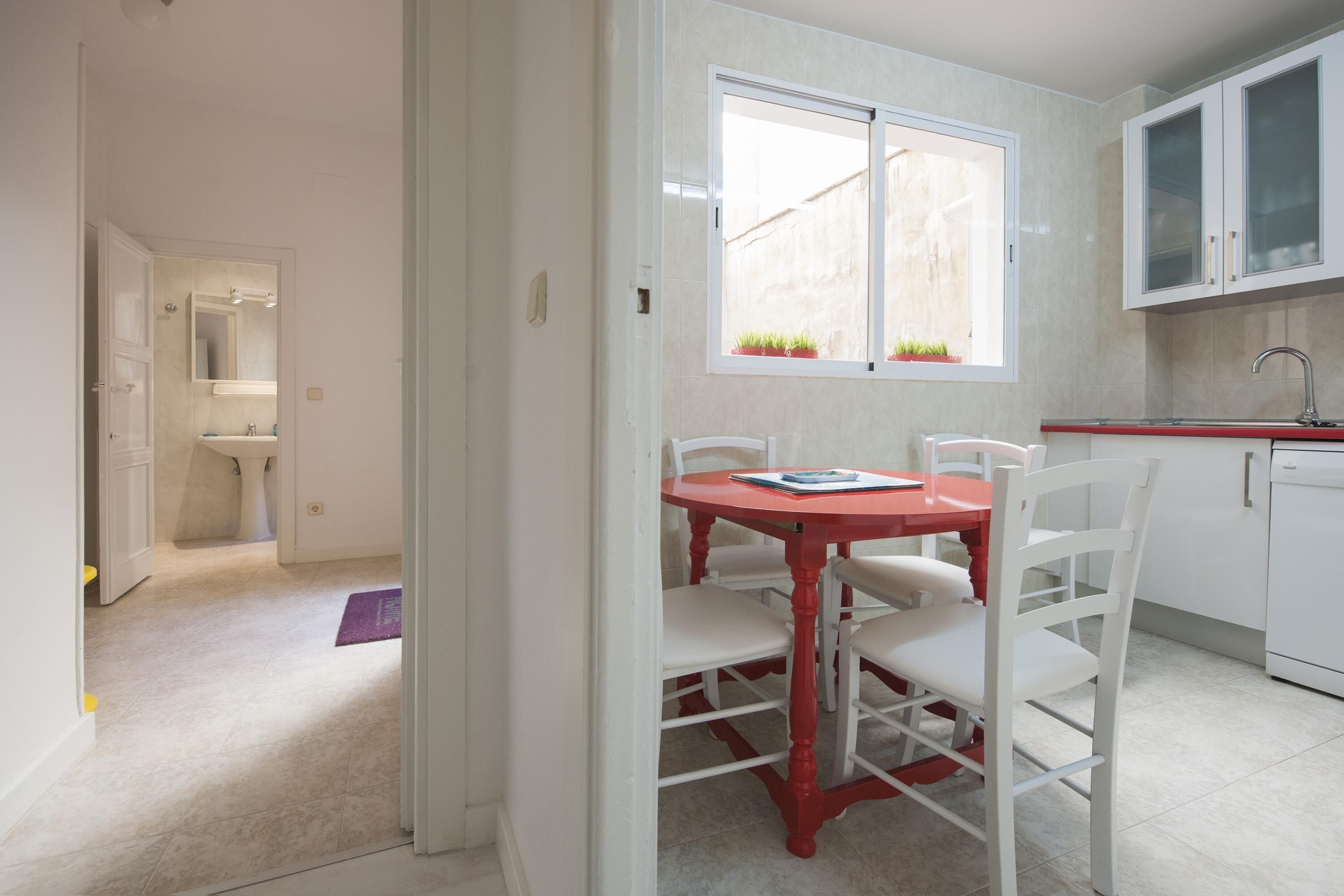 A kitchen with white cabinets, a red table, and chairs. There is a window with plants on the windowsill.